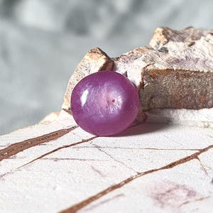 May include: A round, polished, deep purple gemstone with a lustrous surface. The gem is set against a backdrop of textured, light-colored wood and a blurred gray background. The stone's color is a rich, saturated hue.