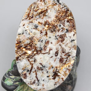 May include: An oval-shaped gemstone with a white base, speckled with black and brown inclusions, and shimmering gold flakes. The polished stone is displayed on a decorative stand with green and black accents, showcasing its natural beauty.