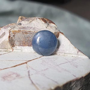 May include: A smooth, round, blue gemstone, possibly a cabochon, sits on a rough, textured, light-colored rock. The gemstone has a glossy surface, reflecting light. The background is blurred, emphasizing the gem's color and shape.