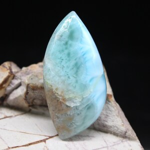 May include: A polished, marquise-shaped larimar gemstone featuring a smooth, glossy surface with a marbled pattern of sky blue, white, and soft tan inclusions. The stone displays a translucent quality with visible internal crystalline textures.
