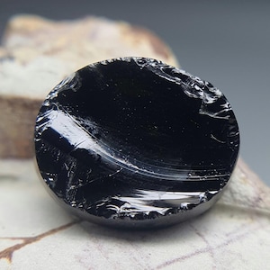 Könnte beinhalten: Ein polierter, runder, schwarzer Obsidianstein. Die Oberfläche reflektiert das Licht und zeigt subtile innere Muster und eine leicht unebene Textur. Der Stein ist vor einem neutralen Hintergrund platziert, der seine dunkle Farbe und natürliche Schönheit hervorhebt.