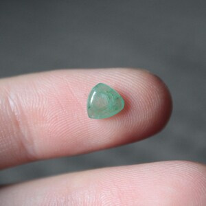 May include: A small, triangular, translucent gemstone with a light green hue. The polished stone has a smooth surface and appears to be held between two fingers. The gemstone is a natural, organic material.