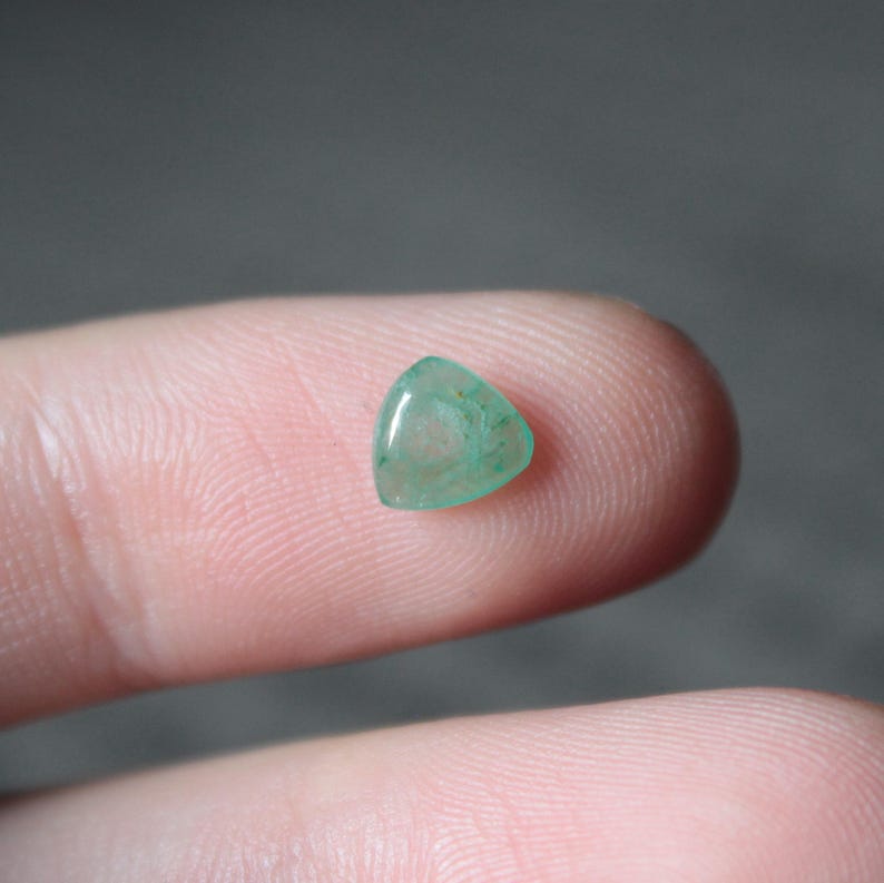 May include: A small, triangular, translucent gemstone with a light green hue. The polished stone has a smooth surface and appears to be held between two fingers. The gemstone is a natural, organic material.