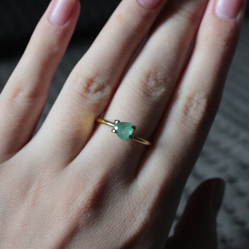 May include: A gold ring featuring a heart-shaped, light green gemstone. The ring's band is thin and simple, with the gemstone held in place by small prongs. The ring is shown on a person's finger.