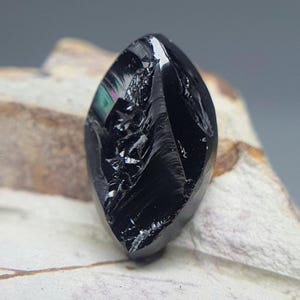 May include: A polished, black gemstone with a teardrop shape. The surface reflects light, revealing subtle iridescent colors. The stone has a rough, textured appearance, with visible fractures and imperfections. The gemstone is displayed on a light-colored, textured surface.