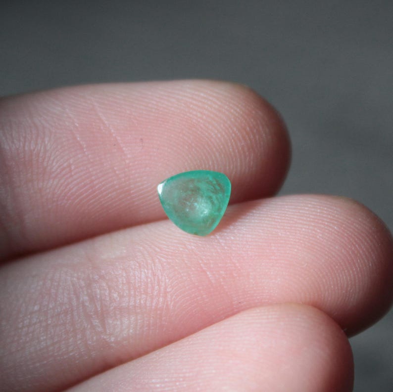 May include: A small, triangular-shaped, translucent emerald gemstone. The gem is a vibrant green color and appears to be faceted. The stone is held between two fingers, with a neutral background.