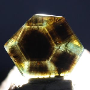 May include: A translucent, hexagonal gemstone with a dark brown center and lighter brown and green hues. The gem is faceted, revealing internal structures and patterns. The stone is set on a light-colored base, with a dark background.