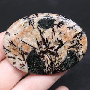 May include: Oval-shaped gemstone with a polished surface. The stone features a beige base color with black, dark green, and gold inclusions. The inclusions are in the form of needle-like crystals and dark patches.