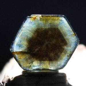 May include: A close-up of a hexagonal gemstone with a translucent, layered appearance. The gem features a central brown core surrounded by a blue and yellow outer layer. The stone is set against a dark background, highlighting its crystalline structure.