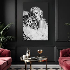 May include: Black and white wall art print featuring a woman with blonde hair and a leopard. The woman is wearing a white feathered shawl and jewelry. The leopard is close to the woman. The print is displayed in a black room with a red sofa and a round table.