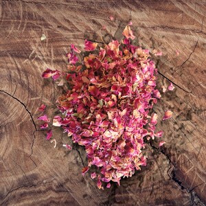 May include: Dried rose petals in shades of pink and red, scattered on a rustic wooden surface.