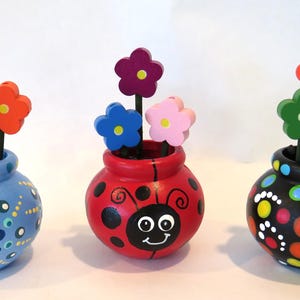 May include: Three decorative ceramic vases, each with a different design and colour scheme. Each vase holds wooden flower cutouts in various colours. The vases are blue, red, and black, with painted details.