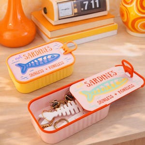 Pode incluir: Dois recipientes de armazenamento retangulares em forma de latas de sardinha. Um é amarelo com um gráfico de peixe azul e o texto not SARDINES SKINLESS + BONELESS. O outro é rosa com um gráfico de peixe amarelo e o texto SARDINES SKINLESS + BONELESS.