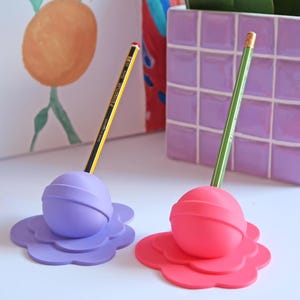 Peut inclure: Deux porte-crayons colorés, un violet et un rose, chacun en forme de fleur avec un dessus sphérique. Un crayon jaune et noir est dans le support violet, et un crayon vert dans le support rose. En arrière-plan, une peinture d'oranges et un vase carrelé violet.