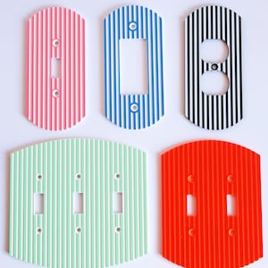 Może przedstawiać: Kolekcja kolorowych, paskowanych osłon na włączniki światła w kolorach różowym, niebieskim, czarnym, zielonym i czerwonym. Osłony mają pionowe paski i różne wycięcia na włączniki i gniazdka. W tle widoczne są dzieła sztuki.