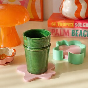 Puede incluir: Un vaso de cerámica verde sobre un posavasos rosa claro con forma de flor. Cerca hay un soporte para posavasos verde menta, una lámpara de hongo naranja, un jarrón ondulado blanco y naranja y un libro titulado St. Tropez Soleil Palm Beach.