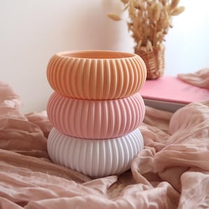 Peut inclure: Trois pots ronds et côtelés empilés, de couleur pêche, rose et blanc. Les pots sont superposés, le pot pêche en haut, le rose au milieu et le blanc en bas. Ils reposent sur un tissu rose.
