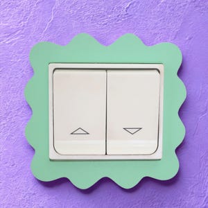 May include: A white double light switch with an upward-pointing triangle on the left button and a downward-pointing triangle on the right button, framed by a light green scalloped border with a wavy edge design.