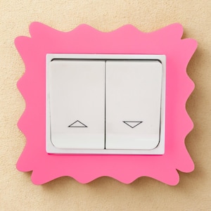 May include: A double light switch featuring two white rectangular rockers with black triangle icons pointing up and down, encased in a bright pink decorative frame with a scalloped, wavy edge design surrounding the central switch mechanism.