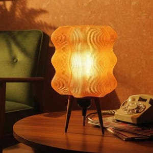 Op de afbeelding: Een oranje tafellamp in retrostijl met een uniek, golvend lampenkapontwerp. De lamp staat op een driepoot met houten poten. De lamp staat op een ronde houten tafel met een draaitelefoon en tijdschriften.