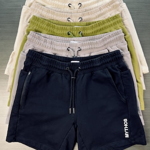 May include: Five pairs of drawstring shorts in different colors, stacked on top of each other. The bottom pair is black with the word "MYTHOS" printed on the right leg.