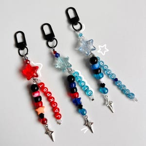 Op de afbeelding: Drie kralen sleutelhangers met zwarte karabijnsluitingen. De eerste heeft een rode ster, zwarte en rode kralen en het woord JACKPOT. De tweede heeft een lichtblauwe ster, blauwe en zwarte kralen en het woord KYRIE. De derde heeft een doorzichtige ster, blauwe en zwarte kralen en het woord THALIA.