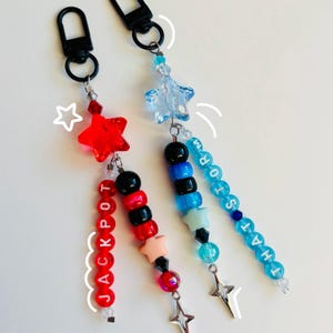 Könnte beinhalten: Zwei Perlen-Schlüsselanhänger mit sternförmigen Anhängern oben. Ein Anhänger hat rote, schwarze und rosa Perlen, die "JACKPOT" buchstabieren, und der andere hat blaue, schwarze und weiße Perlen, die "THAT'S MY STORY" buchstabieren.