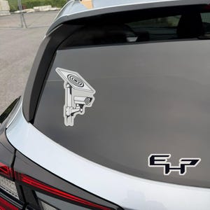May include: A white vinyl decal on a car window featuring a black and white line drawing of a security camera mounted beneath a solar panel. Below this, a separate black and white decal displays the stylized letters E and P in a bold font.