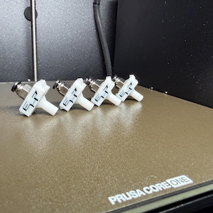 May include: Four white 3D printed plastic components with metallic pneumatic fittings attached. Each component features a T-shaped design with the letters SP printed in black on the front face. These items are arranged in a row on a textured surface.