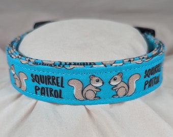 May include: A turquoise blue dog collar with a white background and brown squirrels. The collar has the text "Squirrel Patrol" printed in black.