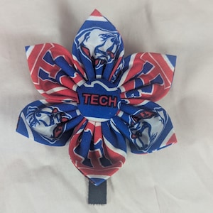 May include: A fabric flower in red, white, and blue, featuring a bulldog graphic and the word "TECH." The flower has six petals and a black strap on the back. The flower is likely a pet accessory.