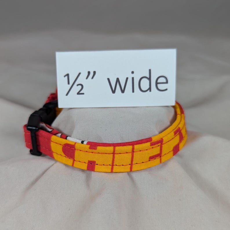 May include: A fabric collar featuring a red and yellow geometric pattern with a black plastic side-release buckle. A white rectangular sign positioned above the collar displays the black text 1/2" wide, indicating the width of the strap.