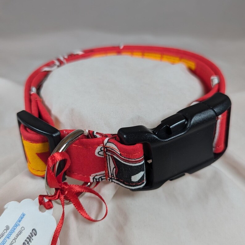 May include: A red fabric pet collar featuring a repeating graphic print of black and white sneakers with yellow accents. The collar includes a black plastic side-release buckle, a silver metal D-ring, and a small red ribbon tag attached.