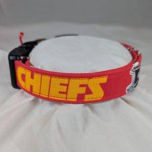 May include: A red fabric pet collar featuring the word CHIEFS printed in bold yellow block letters. The collar includes a black plastic side-release buckle and a metal D-ring for attachment, with visible stitching along the edges of the strap.