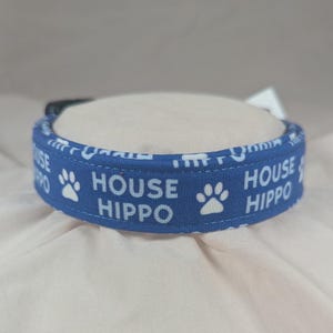 May include: A blue dog collar with the words "HOUSE HIPPO" in white block letters and white paw prints. The collar is made of fabric and has a black buckle.
