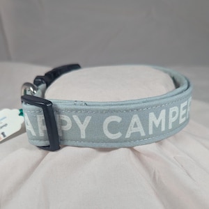 May include: A light blue dog collar with the words "HAPPY CAMPER" printed in white. The collar has a black plastic buckle and metal hardware. The collar is made of a soft fabric.