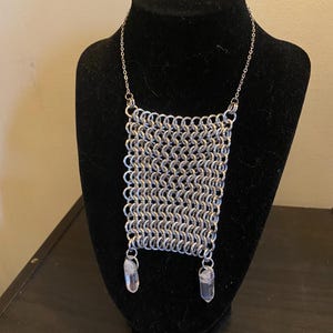 May include: A silver chainmail necklace with a rectangular center piece. Two clear quartz crystal pendants hang from the bottom of the chainmail. The necklace is displayed on a black mannequin.