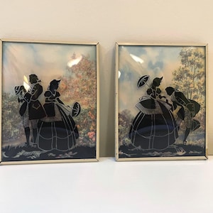 May include: Two framed silhouette art pieces. Each features a black silhouette of a couple in period clothing against a colorful, blurred background. One couple is standing, the other is bowing. The frames are gold-toned.