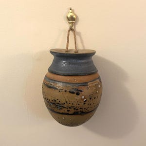 May include: A wall-mounted ceramic vessel with a rounded body and a flared rim. The piece features a blue glazed neck and a tan lower section with dark brown and black speckled accents. It is suspended by a jute cord from a gold-toned metal hook.