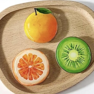 May include: A wooden tray displays three fruit-shaped decorations: an orange, a grapefruit slice, and a kiwi slice. The orange is yellow-orange with a green leaf. The grapefruit slice is white and orange. The kiwi slice is green with a yellow centre.