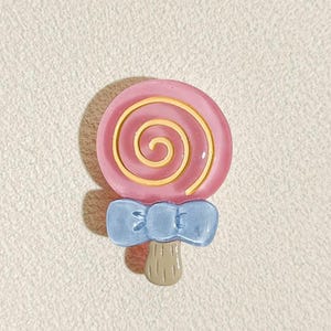 Puede incluir: Una piruleta de espiral rosa y amarilla con un lazo azul y un palo marrón.