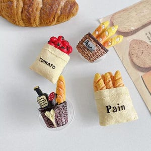 May include: Four miniature fabric bags with different food items inside. The bags are beige and have the words "Pain" and "Tomato" printed on them. The food items include a bunch of red tomatoes, a baguette, and a loaf of bread.