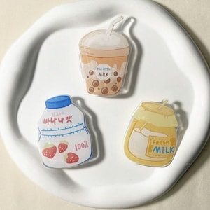 May include: Three colourful cartoon-style drink charms. A brown and white boba tea charm with the text "Tea with Milk", a white milk carton charm with the text "100%" and a strawberry graphic, and a yellow glass jar charm with the text "Fresh Milk".