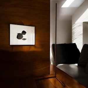 May include: A modern interior scene featuring a framed abstract artwork with black ink blotches on a white background. The artwork hangs on a dark wood-paneled wall. A tall, slender metal floor lamp stands near a dark-colored sofa.