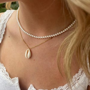 May include: A layered necklace featuring a delicate pearl strand and a gold chain. The lower chain holds a white seashell pendant. The jewelry is displayed against a white ribbed top with lace trim, creating a simple, elegant look.