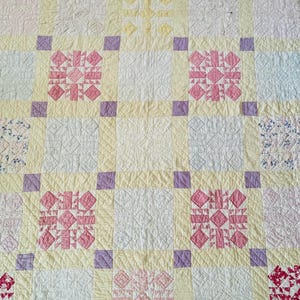 May include: A handmade quilt with a patchwork design. The quilt features a light yellow background with squares of white, pink, and patterned fabrics. Pink and red floral patterns are centered in some squares, with purple accents.