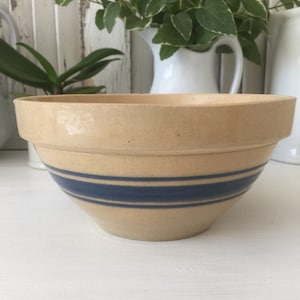 May include: A beige ceramic mixing bowl featuring a wide rim and two horizontal dark blue stripes encircling the lower exterior. The bowl has a smooth, matte finish and a rounded base, designed for kitchen use and food preparation tasks.