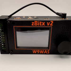 May include: A black zBitx v2 radio transceiver featuring a central rectangular screen, a large circular tuning knob on the right, and a side-mounted cooling vent. The front panel displays the text zBitx v2 and W9WAS in orange lettering.