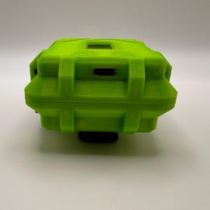 May include: A bright green, rectangular, rugged case with a textured surface. The case has a small rectangular opening on top and a USB port on the side. The case is mounted on a black base.