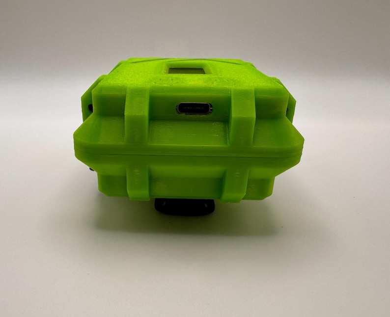 May include: A bright green, rectangular, rugged case with a textured surface. The case has a small rectangular opening on top and a USB port on the side. The case is mounted on a black base.